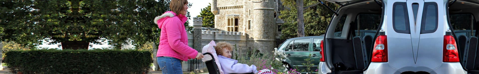 Photo of a daughter caring for her disabled mother by assisting her into the back of a wheelchair access vehicle after spending a lovely day out visiting a castle for cream teas